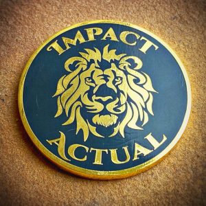 Front of a gold and black challenge coin with a detailed lion face design and the text “Impact Actual” around the edge.